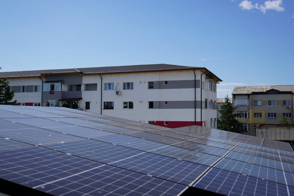 Rooftop solar panels in the foreground with white and gray apartment buildings under a clear blue sky in the background.