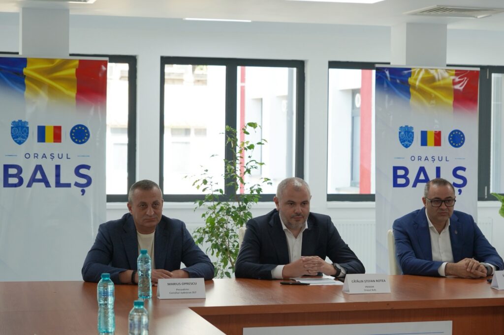Three men in suits sit at a panel table during a city event, with banners reading 'Orașul Balș' and flag imagery in the background.