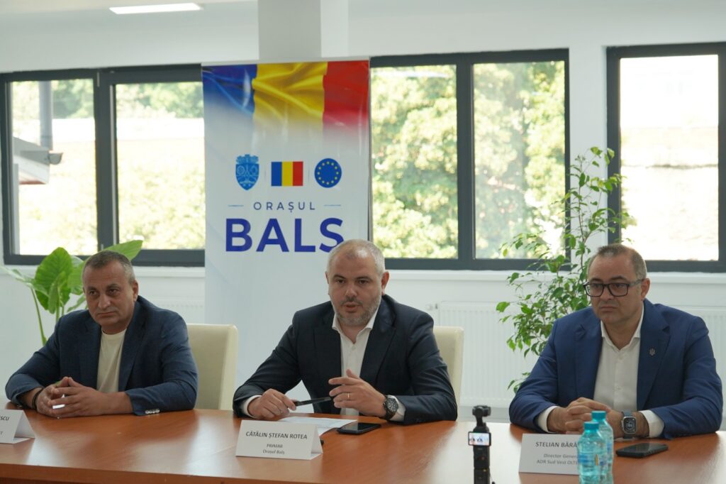 Three men in suits sit at a conference table with a 'ORAȘUL BALS' banner behind them.