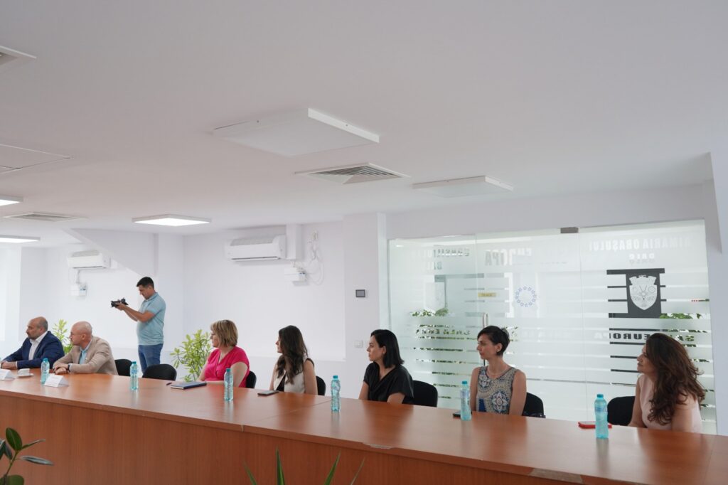 Group of professionals seated along a long conference table in a bright office, while a man takes a photo with a camera nearby.