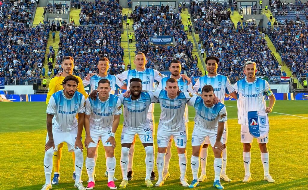 Soccer team in light blue and white striped uniforms posing for a group photo on the pitch with fans in the stands behind them.