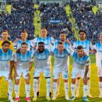 Soccer team in light blue and white striped uniforms posing for a group photo on the pitch with fans in the stands behind them.