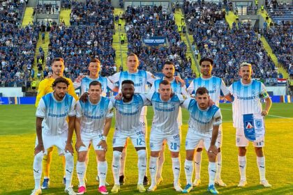 Soccer team in light blue and white striped uniforms posing for a group photo on the pitch with fans in the stands behind them.