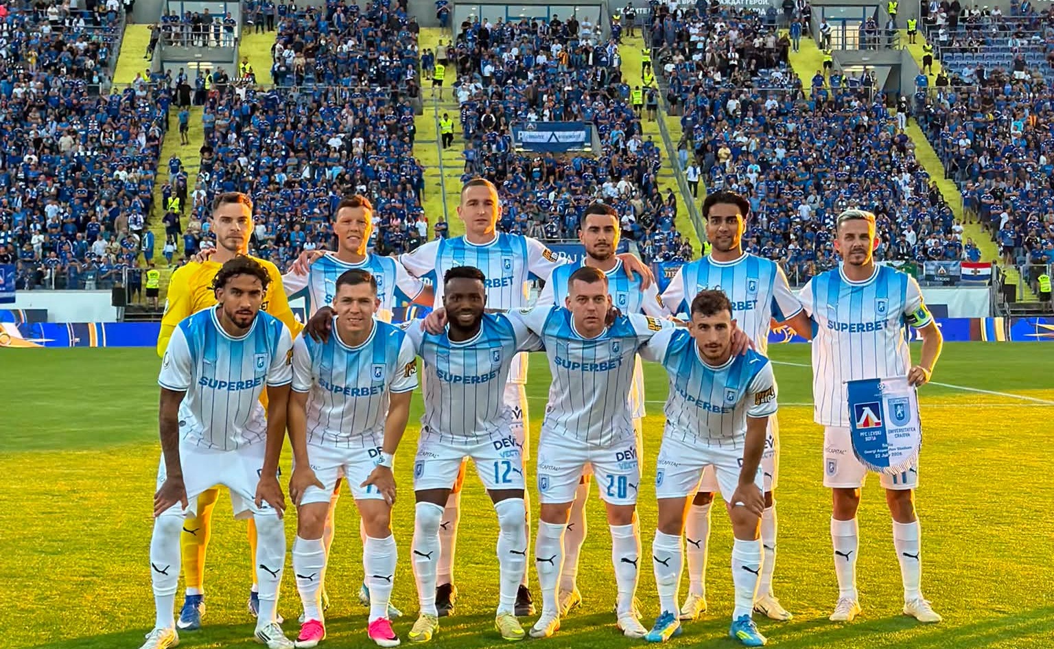 Soccer team in light blue and white striped uniforms posing for a group photo on the pitch with fans in the stands behind them.