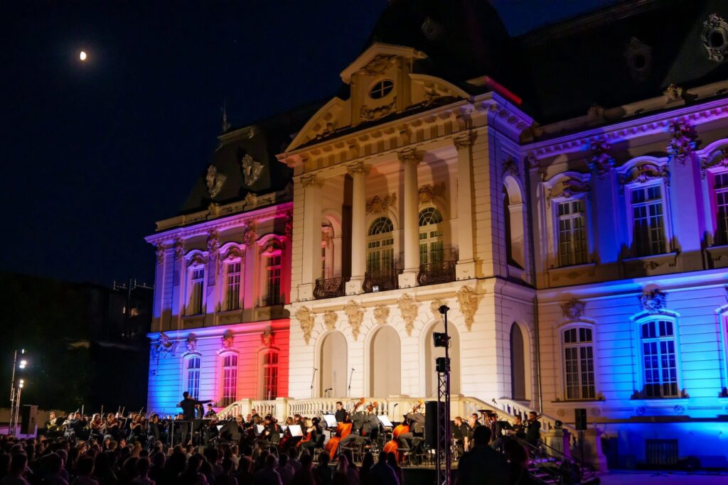 Nighttime outdoor orchestra performance in front of a lit historic building with red, white, and blue lights.