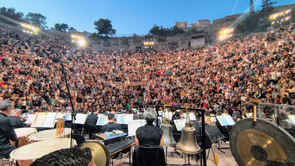 Public Odeon Atena 2026 - Jurnalul Olteniei – Știri din Craiova și Oltenia Orchestra performing on stage in an ancient open-air amphitheater, with a large audience in stone-tiered seating beyond dancers and singers panning the crowd.
