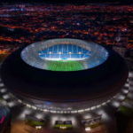 Aerial night view of a large circular stadium with a brightly lit green and blue field inside the ring-shaped arena amid a cityscape.