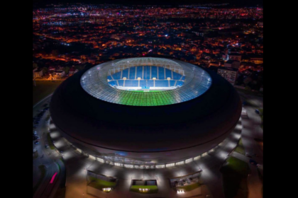 Aerial night view of a large circular stadium with a brightly lit green and blue field inside the ring-shaped arena amid a cityscape.
