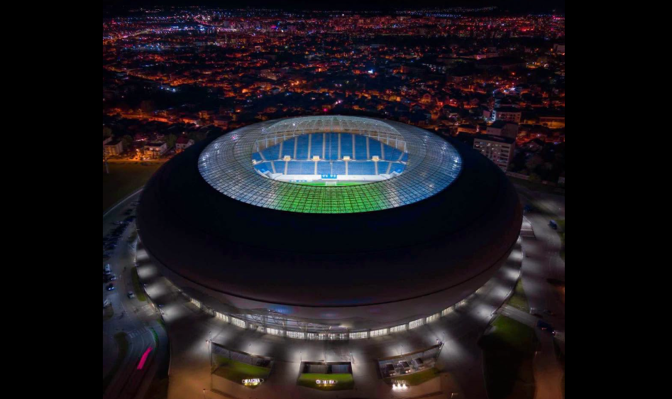 Aerial night view of a large circular stadium with a brightly lit green and blue field inside the ring-shaped arena amid a cityscape.