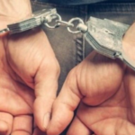 Person with hands cuffed behind their back, wearing metal restraints connected by a chain behind them