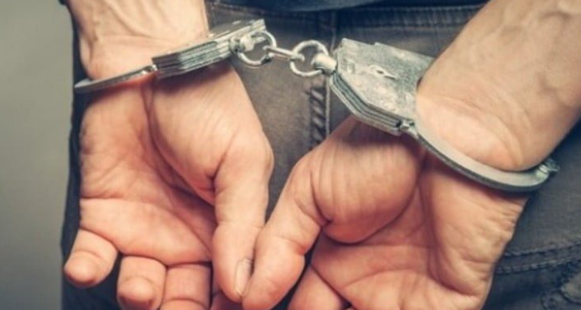 Person with hands cuffed behind their back, wearing metal restraints connected by a chain behind them