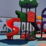 Colorful playground structure with red spiral slide, green and purple ramps, and a bright red straight slide in a sunny outdoor park area.