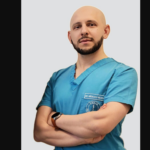 Male doctor in teal scrubs with arms crossed, wearing a smartwatch against a light gray backdrop