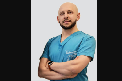 Male doctor in teal scrubs with arms crossed, wearing a smartwatch against a light gray backdrop