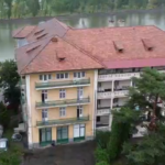 A row of beige apartment buildings with red-tiled roofs beside a river, surrounded by trees and a flooded street below.