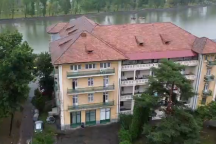 A row of beige apartment buildings with red-tiled roofs beside a river, surrounded by trees and a flooded street below.