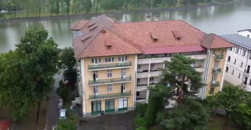 A row of beige apartment buildings with red-tiled roofs beside a river, surrounded by trees and a flooded street below.