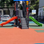 Playground with orange and green slides attached to a central climbing structure on a red and green safety surface, in an urban area.