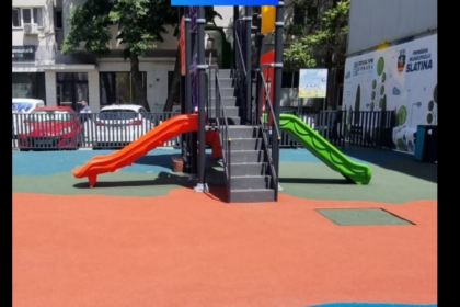 Playground with orange and green slides attached to a central climbing structure on a red and green safety surface, in an urban area.