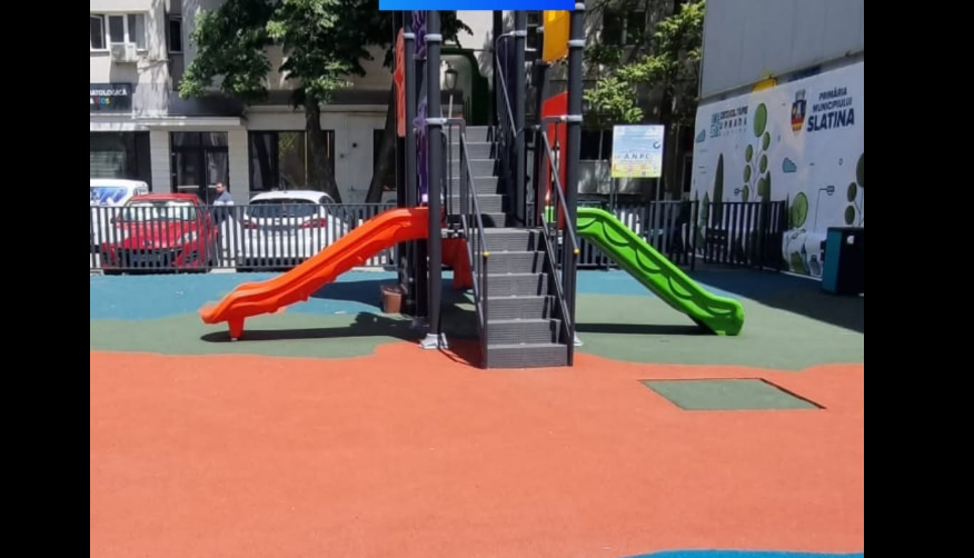 Screenshot 2026-07-20 092413 - Jurnalul Olteniei – Știri din Craiova și Oltenia Playground with orange and green slides attached to a central climbing structure on a red and green safety surface, in an urban area.