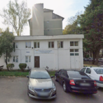 White, two-story office building with a sign for the Pensioners’ Association, parked cars in front, and trees on both sides of a small lot.
