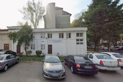White, two-story office building with a sign for the Pensioners’ Association, parked cars in front, and trees on both sides of a small lot.