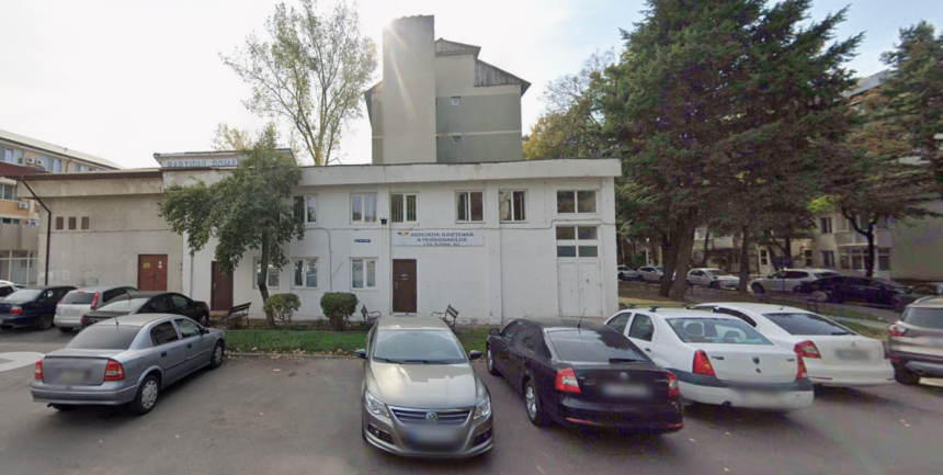 White, two-story office building with a sign for the Pensioners’ Association, parked cars in front, and trees on both sides of a small lot.