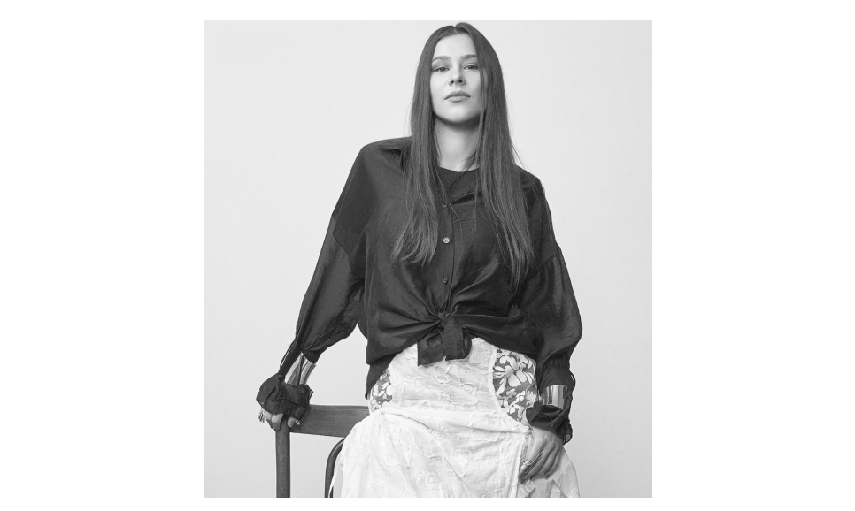 Screenshot 2026-07-21 094632 - Jurnalul Olteniei – Știri din Craiova și Oltenia Woman seated, wearing a knotted black blouse and a floral lace skirt, looking at the camera in a studio portrait.