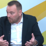 Man in a dark blazer and white shirt talks, using hand gestures, seated at a glass table in a TV studio backdrop with yellow patterns.