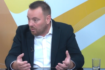 Man in a dark blazer and white shirt talks, using hand gestures, seated at a glass table in a TV studio backdrop with yellow patterns.