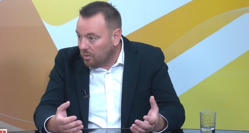 Man in a dark blazer and white shirt talks, using hand gestures, seated at a glass table in a TV studio backdrop with yellow patterns.