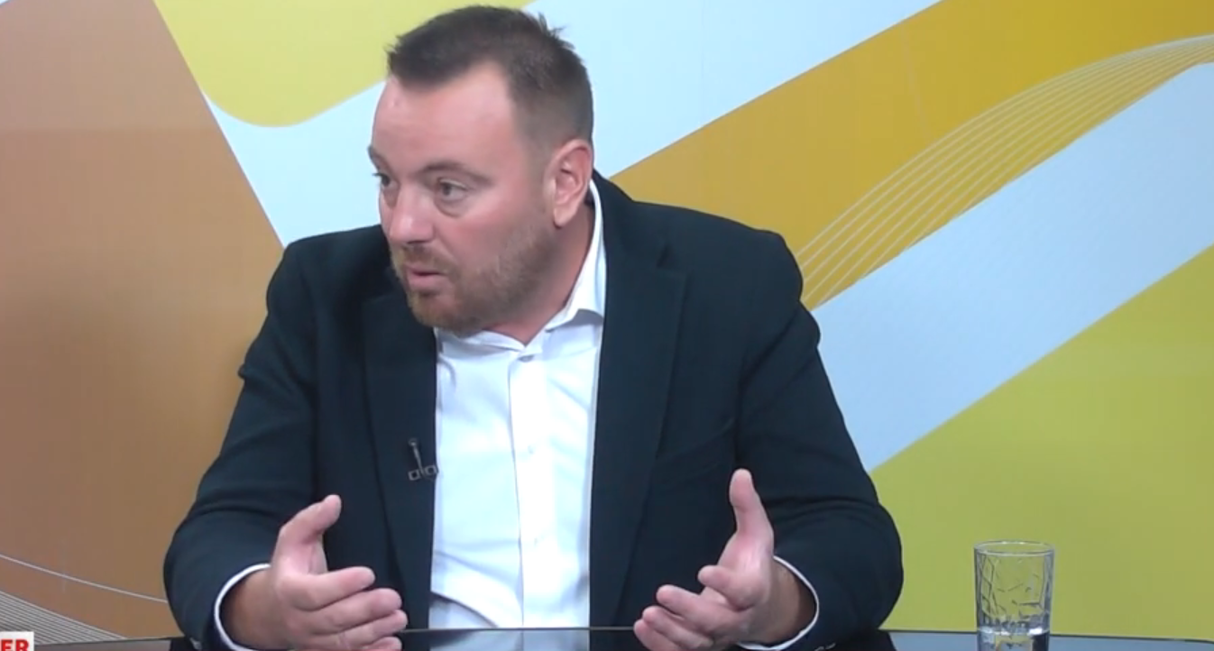 Man in a dark blazer and white shirt talks, using hand gestures, seated at a glass table in a TV studio backdrop with yellow patterns.