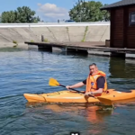 Person wearing an orange life jacket paddling a yellow kayak on calm water, with a wooden boathouse and dock in the background.