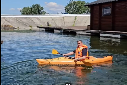 Person wearing an orange life jacket paddling a yellow kayak on calm water, with a wooden boathouse and dock in the background.