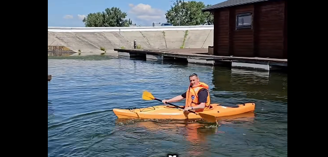 Person wearing an orange life jacket paddling a yellow kayak on calm water, with a wooden boathouse and dock in the background.