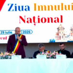 Man wearing a Romanian sash speaks at a podium during Ziua Imnului Național ceremony; two officials sit at a flowered table behind him with a large banner reading 'Ziua Imnului Național'.
