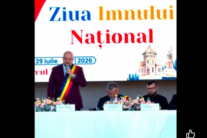Man wearing a Romanian sash speaks at a podium during Ziua Imnului Național ceremony; two officials sit at a flowered table behind him with a large banner reading 'Ziua Imnului Național'.