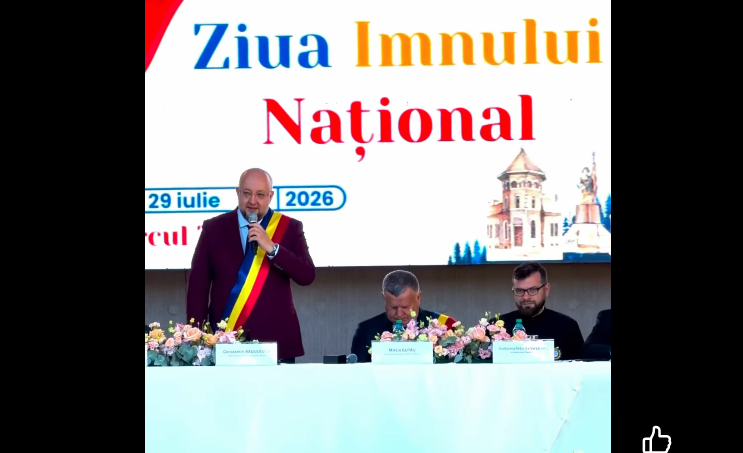 Man wearing a Romanian sash speaks at a podium during Ziua Imnului Național ceremony; two officials sit at a flowered table behind him with a large banner reading 'Ziua Imnului Național'.