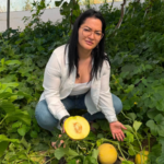 Woman kneeling in a melon patch, presenting a halved cantaloupe with a whole melon nearby to the camera.