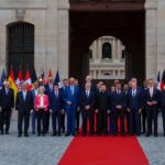 Group of formally dressed officials standing on a red carpet in front of a historic stone building with flags behind them.