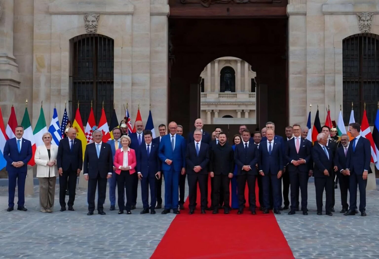 Group of formally dressed officials standing on a red carpet in front of a historic stone building with flags behind them.