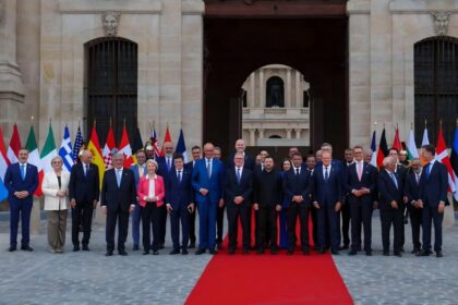 Group of formally dressed officials standing on a red carpet in front of a historic stone building with flags behind them.