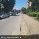 Urban street in Târgu Jiu, Romania, with parked cars along the left curb, a sidewalk on the right, and trees and buildings under a clear blue sky.