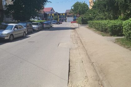 Urban street in Târgu Jiu, Romania, with parked cars along the left curb, a sidewalk on the right, and trees and buildings under a clear blue sky.