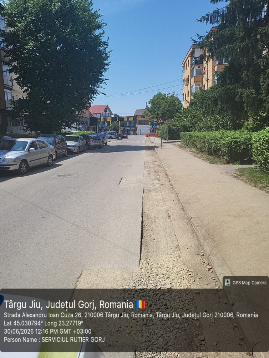 Urban street in Târgu Jiu, Romania, with parked cars along the left curb, a sidewalk on the right, and trees and buildings under a clear blue sky.