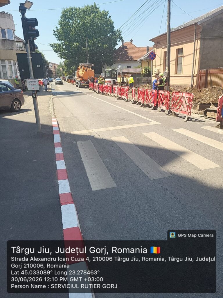 WhatsApp Image 2026-07-01 at 111445 - Jurnalul Olteniei – Știri din Craiova și Oltenia Roadwork scene on a sunny street in Târgu Jiu, Romania, with red-and-white barriers lining the sidewalk and construction vehicles in the road.