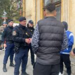 Police officers in tactical gear speak with a man in a black puffer jacket on a city street beside a weathered yellow building.