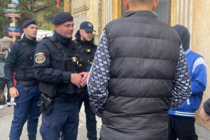 Police officers in tactical gear speak with a man in a black puffer jacket on a city street beside a weathered yellow building.