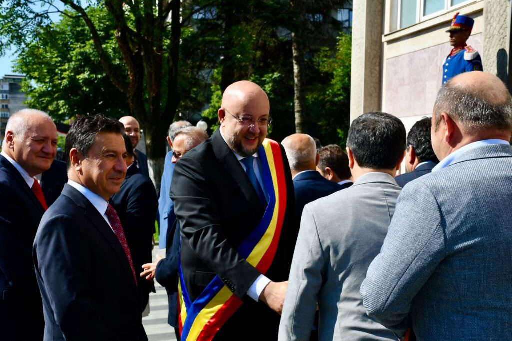 Bald man in a suit wearing a Romanian tricolor sash greets people at an outdoor formal event.