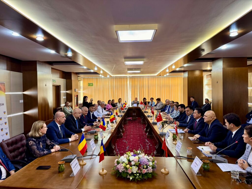 International meeting with delegates seated around a long U-shaped wooden table, flags of multiple nations on the desk, and a central floral centerpiece in a formal conference room.